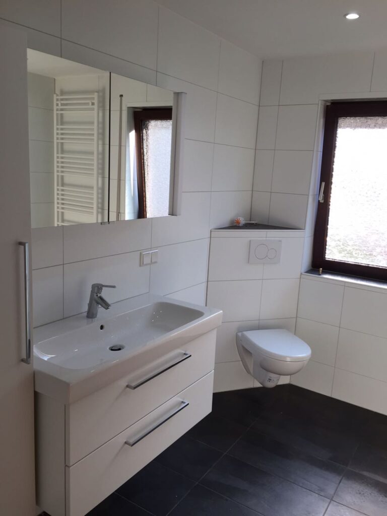 Weißes saniertes Badezimmer mit Waschtisch, Spiegelschrank, wandhängendem WC und dunklem Fliesenboden, Badsanierung