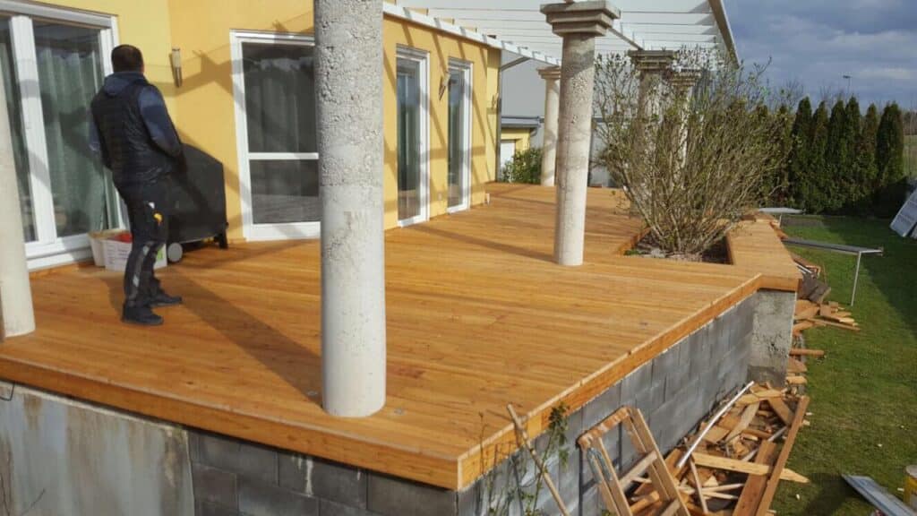 Handwerker verlegt Holzterrasse auf Betonunterbau mit Stützen und Holzresten, Bodenverlegung