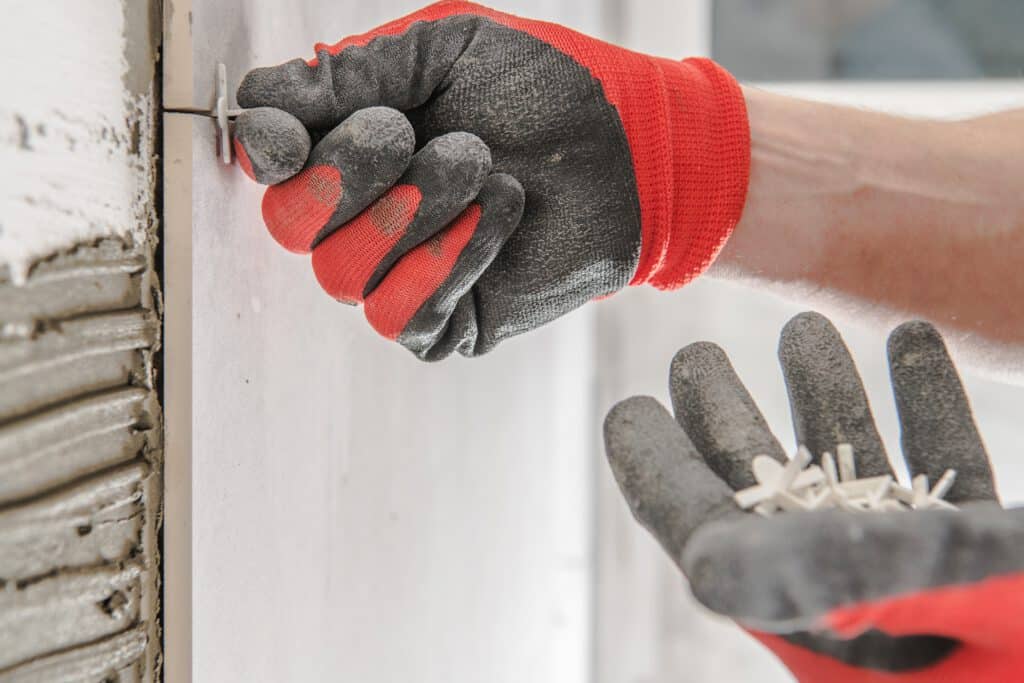 Hand mit Handschuhen setzt Fliesenkreuz zwischen Wandfliesen bei Badsanierung und Fliesenarbeiten